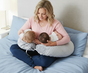 Woman breastfeeding two babies using a nursing pillow on a bed.