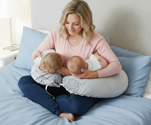 Woman breastfeeding two babies using a nursing pillow on a bed.