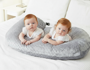 Two babies lying on a gray baby pillow on a white bed.