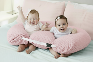Two babies sitting on a gray body pillow on a bed.