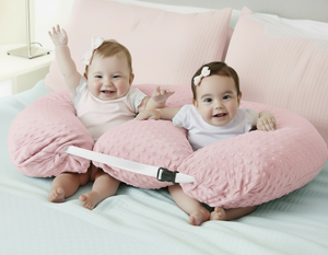 Two babies sitting on a gray body pillow on a bed.