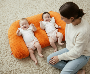 Two babies lying on a gray U-shaped pillow on a dark gray carpet.