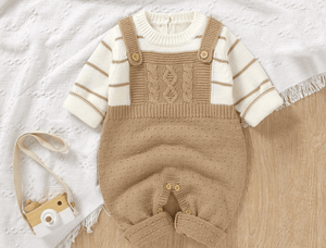 Baby outfit with beige knitted overalls and striped shirt on a light wooden surface.