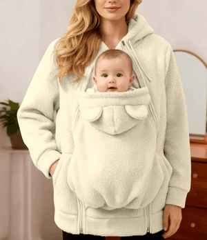 Person wearing a cream-colored baby carrier with a baby inside, standing indoors.
