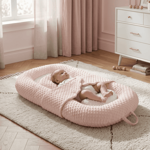 Baby lying on a pink circular mat in a nursery with white furniture and curtains.