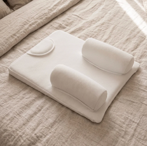 A white ergonomic baby support pillow set with a contoured mattress and two cylindrical head/neck pillows, styled on a beige linen bed.