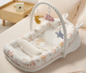 A baby bassinet with an attached white arch activity toy, decorated with a soft pastel floral and animal print. The bassinet is styled on a beige bed with a wooden side table in the background.