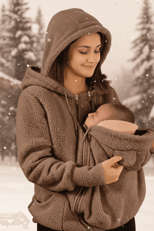 A woman wearing a brown sherpa fleece zip-up hoodie with a baby carrier pouch holding a sleeping newborn baby, set against a snowy outdoor background with a heat icon overlay.