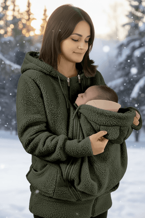 A woman wearing an olive green fleece hoodie with a built-in baby carrier pocket holding a baby, set against a snowy outdoor background with a flame graphic indicating warmth.