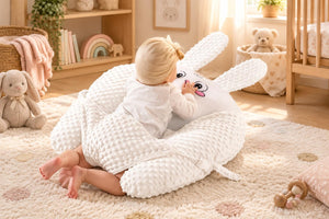 A baby wearing a white outfit and a beige bonnet lies on a white textured bunny-shaped baby support pillow on a beige bed.