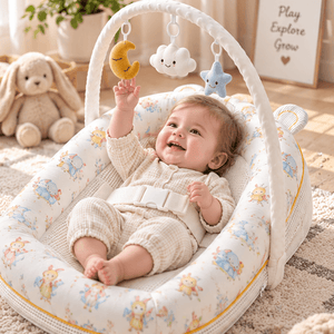 A baby lies in a padded bassinet with a white arch and hanging toys, photographed in a softly styled nursery. The bassinet fabric is cream with a small pastel animal print, and the baby is wearing a matching cream outfit.