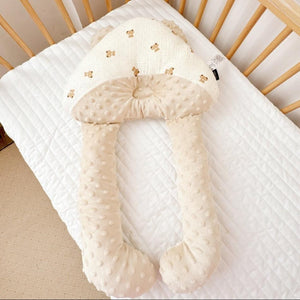 A beige, C-shaped newborn baby head shaping pillow with a velvet finish, placed on a white textured surface.