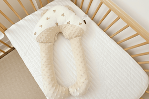 A beige, C-shaped newborn baby head shaping pillow with a velvet finish, placed on a white textured surface.