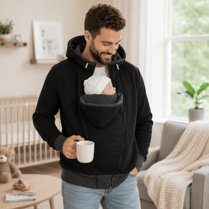 A man wearing a black baby carrier hoodie with a baby tucked inside, standing in a bright living room and holding a white mug.
