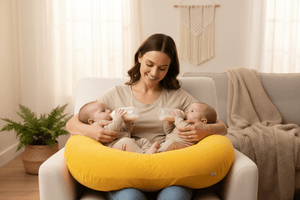Woman holding two babies with a yellow nursing pillow in a cozy living room.
