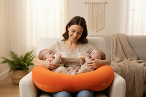 Woman holding two babies with a yellow nursing pillow in a cozy living room.