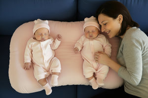 Two babies in matching outfits with toys on a green cushion, accompanied by an adult.
