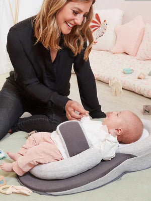 A smiling woman is attending to a newborn baby lying in an inclined anti-reflux baby lounger with safety straps. The lounger is placed on a bed, and the baby is wearing light-colored clothes.
