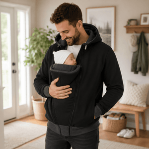 A man wearing a black zip-up hoodie with a baby carrier insert holds a sleeping newborn baby in a home setting.
