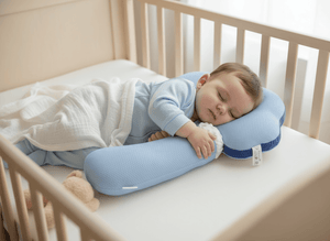 Baby sleeping on a U-shaped pillow in a crib with a teddy bear beside.