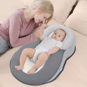 Baby lying on a gray and white baby lounger pillow with a woman sitting beside it on a beige couch.