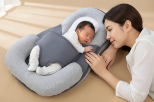 Baby sleeping in a gray and white baby lounger pillow with a woman beside it on a beige surface.