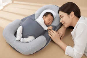 Baby sleeping in a gray and white baby lounger pillow with a woman beside it on a beige surface.