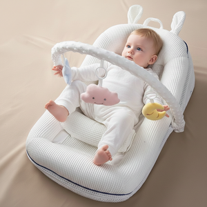 Baby lying on a white textured cushion with toys, sitting up with support from a white crib safety bar.