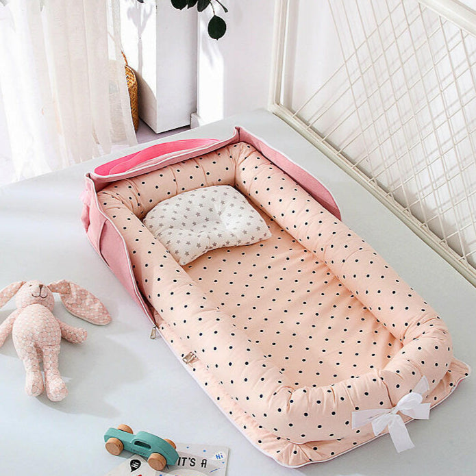 A portable Travel Baby Sleeper Bed with pink and white polka dot pattern, set up on the floor with a white pillow inside it, next to a teddy bear and a toy car.