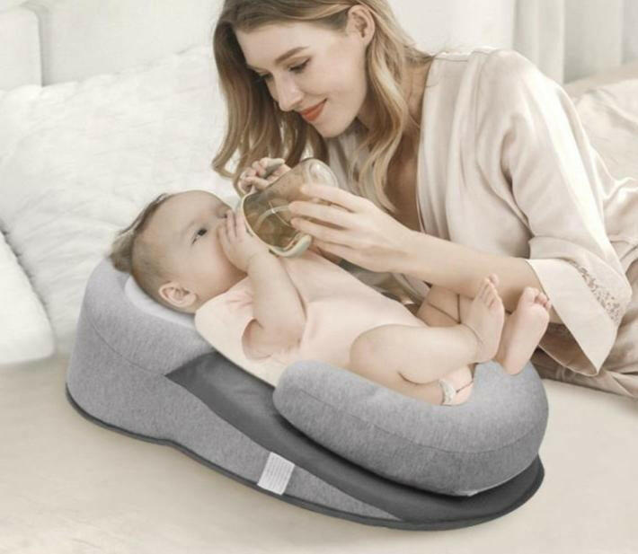 A baby lying on an inclined Anti-Reflux Baby Feeding & Breastfeeding Support Pillow with a woman sitting next to it, holding the baby.