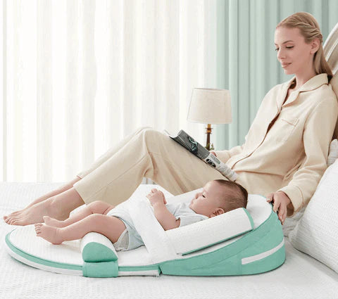 A baby lying on a green and white anti-reflux and anti-roll baby lounger bed while a woman sits next to it reading a book.