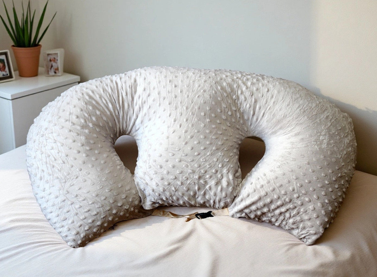White U-shaped pillow on a bed in a bedroom setting with a plant and photos in the background.