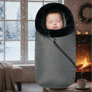 Baby sleeping in a gray swaddle with a black hood in a cozy room with a snowy window view.