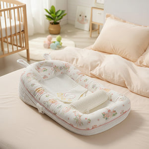A baby bassinet with a white floral print and a matching rolled blanket, styled on a beige bed with pillows and a wooden side table in the background. A pink label in the top left corner reads 'Garden Afternoon Tea'.