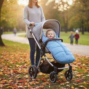 A baby in a light blue quilted footmuff sits in a gray stroller being pushed by an adult woman through a park path covered with fallen leaves.