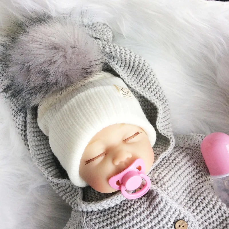 A baby wearing a gray knitted winter hat with a faux fur brush on a white furry background.