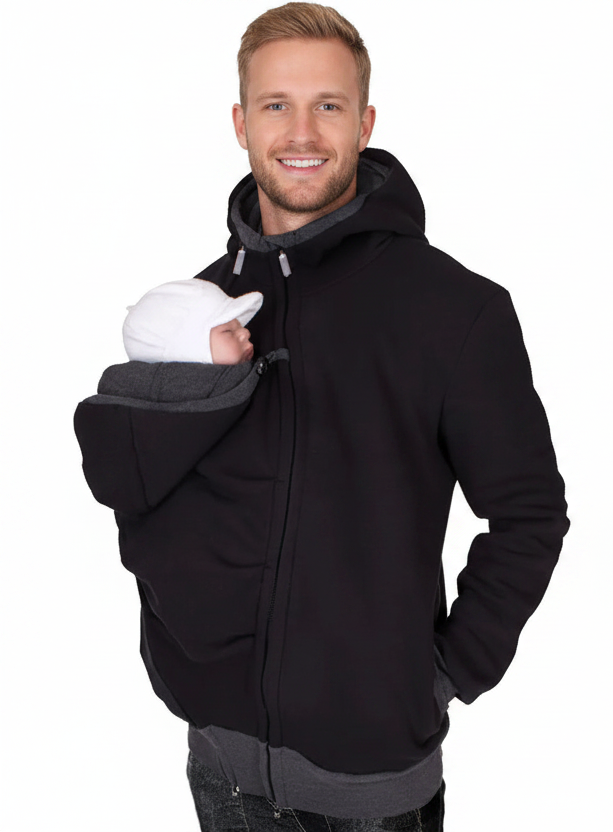 A smiling man wearing a black hooded jacket with a gray inner hood and pocket, holding a baby wrapped in a white blanket against his chest, set against a plain white background.