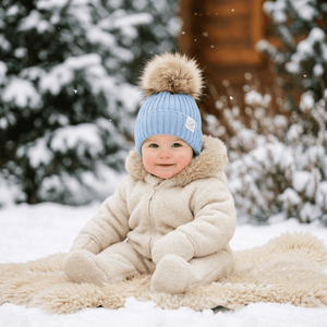 A baby sitting in the snow wearing a cream-colored winter snowsuit and a light blue knit beanie with a large tan faux-fur pom-pom. The background shows snow-covered evergreen trees and a wooden cabin.