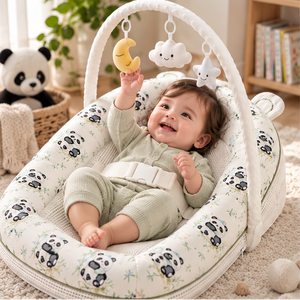 A baby in a pale green knit outfit sits in a white padded bassinet with a panda print, holding a yellow crescent moon toy. The bassinet has an arch overhead with hanging cloud and star toys.