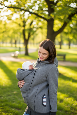 A woman in a gray baby carrier hoodie holds a baby in a white hat in a sunny park with green trees.