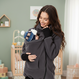A woman in a gray baby carrier jacket holding a baby in a nursery setting.