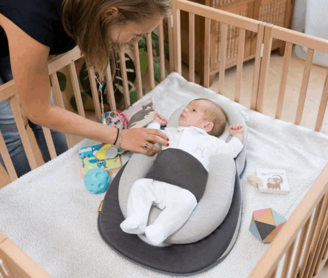 A newborn baby lying in an anti-reflux elevated baby lounger with a woman attending to the baby. The lounger is placed in a crib with toys around and has a gray color scheme.