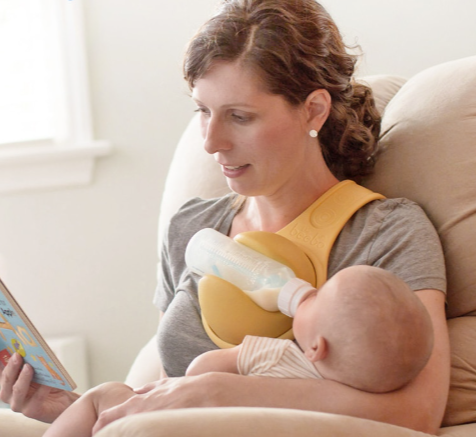A person is seated while wearing a yellow hands-free baby bottle holder in which a baby bottle is placed, allowing the wearer to hold a baby and read a book simultaneously.