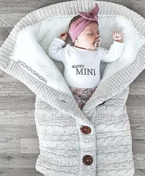 A beige-colored baby sleeping bag with a cable knit pattern, featuring a hood and button closures, with an infant inside.