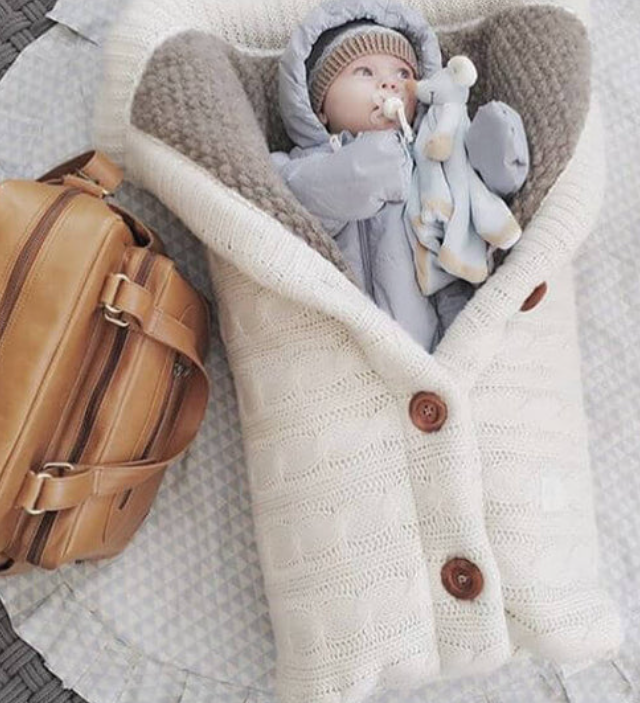 A beige-colored baby sleeping bag with a cable knit pattern, featuring a hood and button closures, with an infant inside.