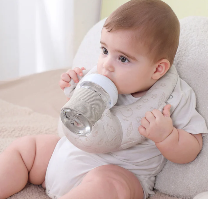 A baby is sitting on a soft surface, wearing a light-colored onesie, and using a hands-free feeding pillow to hold a bottle.