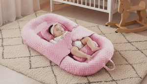 Baby in a pink bassinet in a nursery with a crib and toys in the background.
