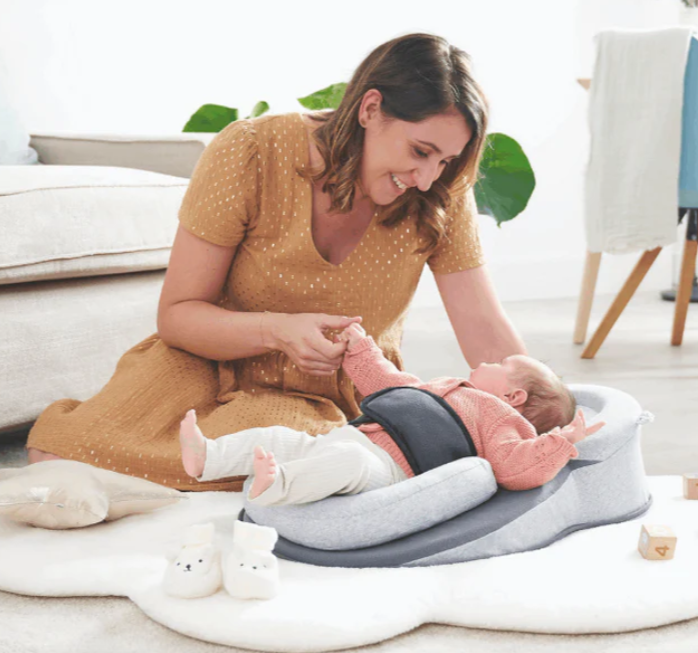 A baby lying in a gray inclined reflux relief baby lounger with a woman sitting next to it, engaging with the baby.