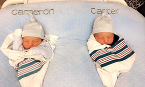 Two newborn babies swaddled in striped blankets with 'Cameron' and 'Carter' on a blue pillow.