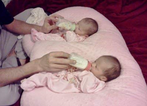 Two babies lying on a pink cushion, being fed with bottles.
